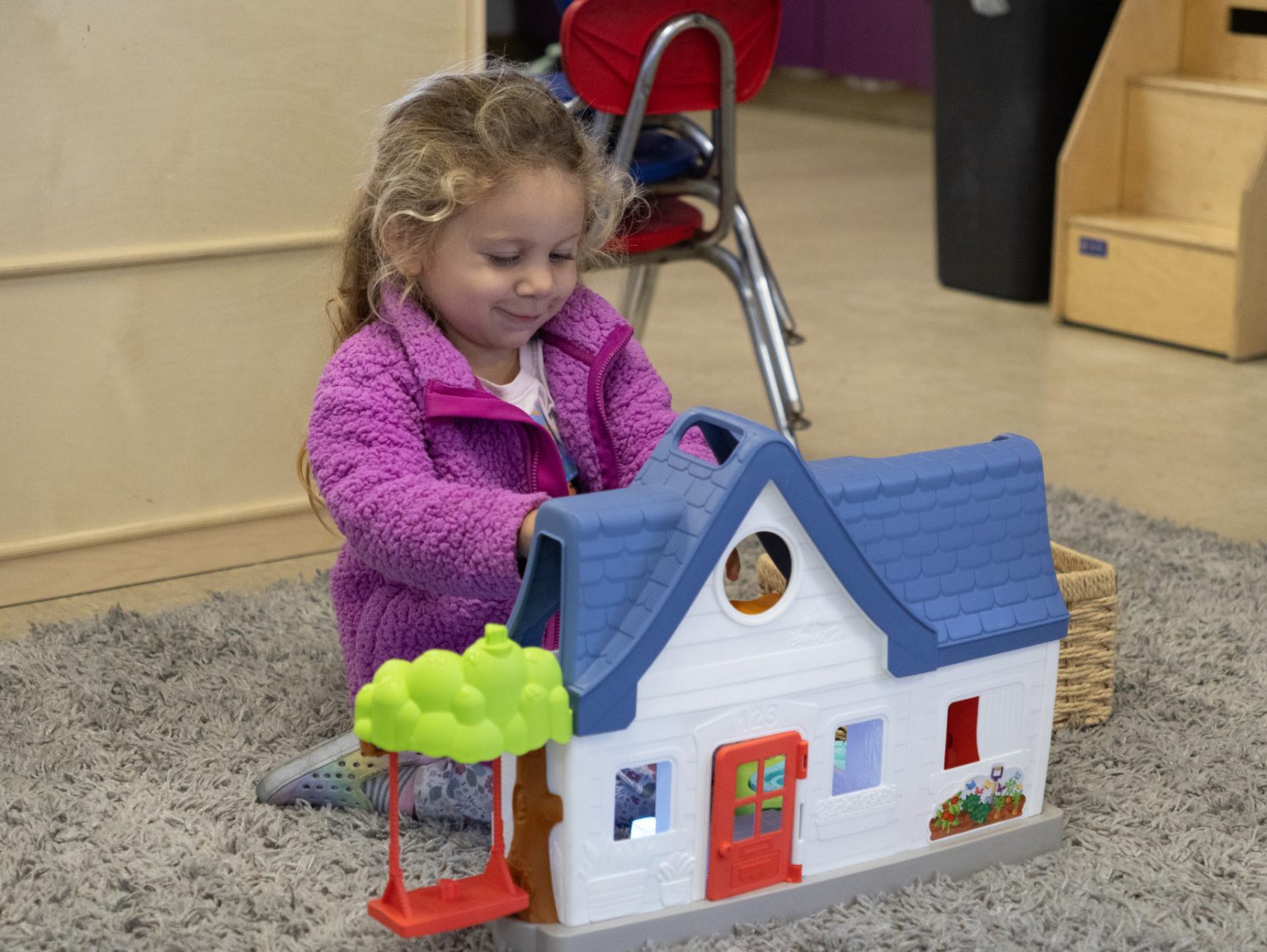 A preschool-aged girl sits on a rug playing with a toy house