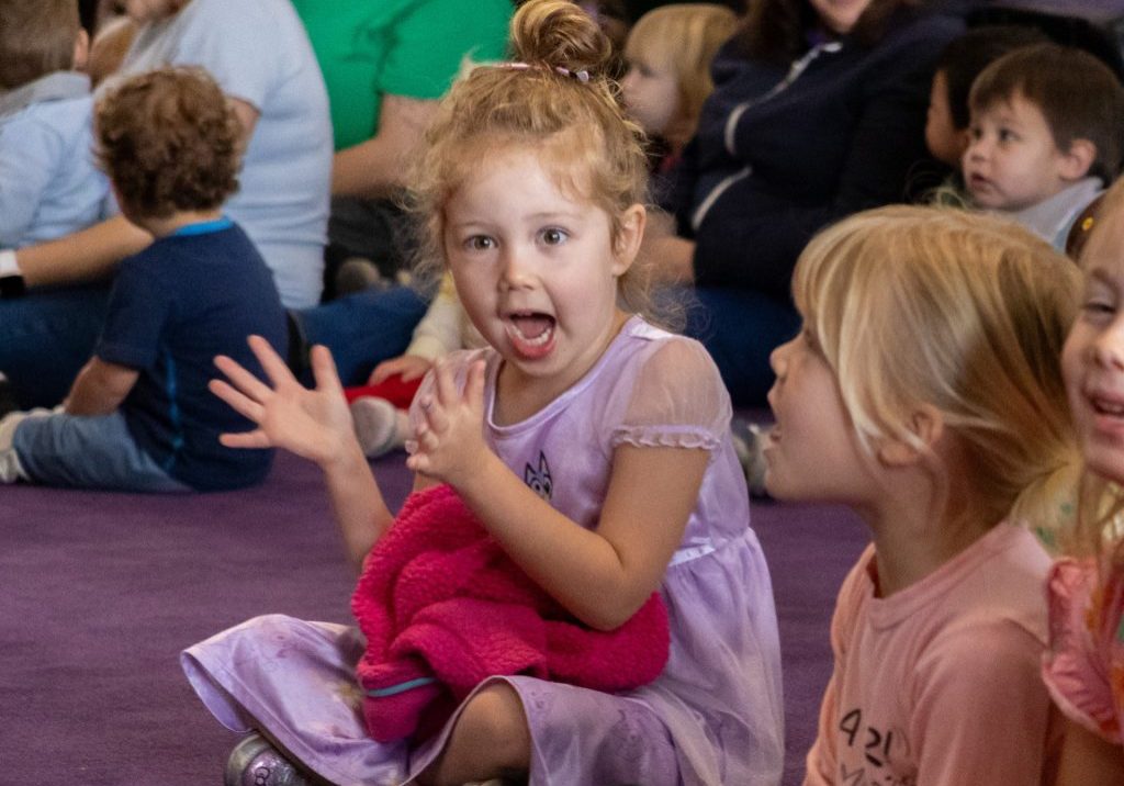 An excited girl, clapping while seated cross-legged on the carpet during a preschool musical performance